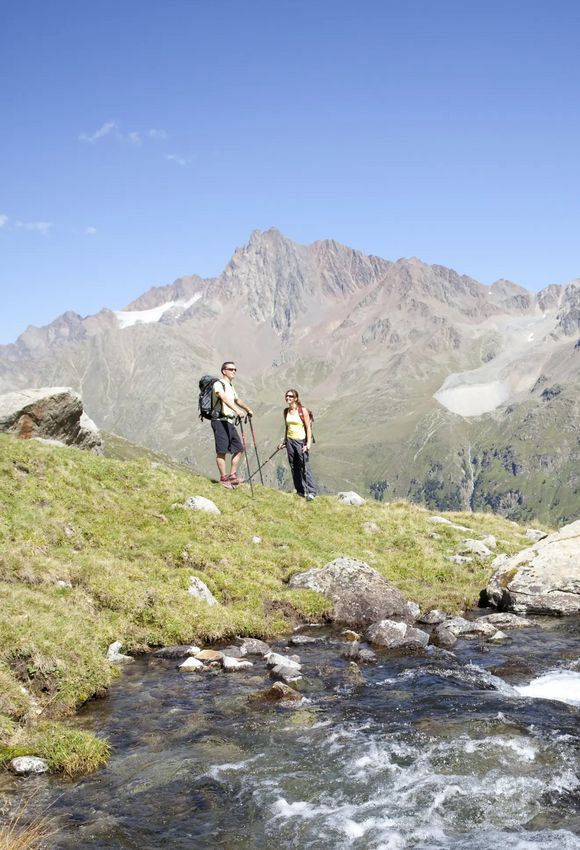 Zwei Wanderer stehen neben einem klaren Gebirgsbach, umgeben von grünen Wiesen und mächtigen Berggipfeln. Der Bach sprudelt über Steine ins Tal.