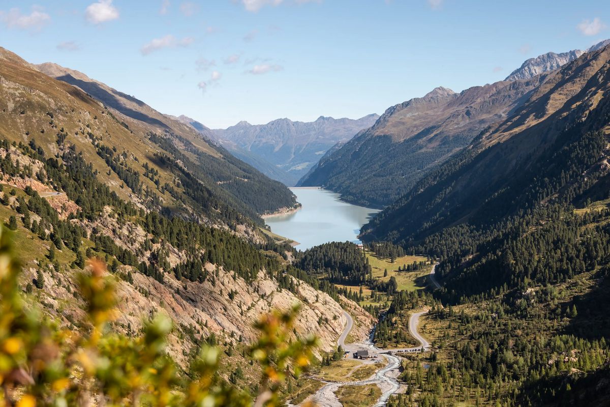 Panoramablick auf den Gepatsch-Stausee im Kaunertal. Der türkisfarbene See liegt eingebettet zwischen bewaldeten Berghängen, die sich bis zum Horizont ziehen. Eine gewundene Straße führt durch das Tal – Weite, Natur, Ruhe.