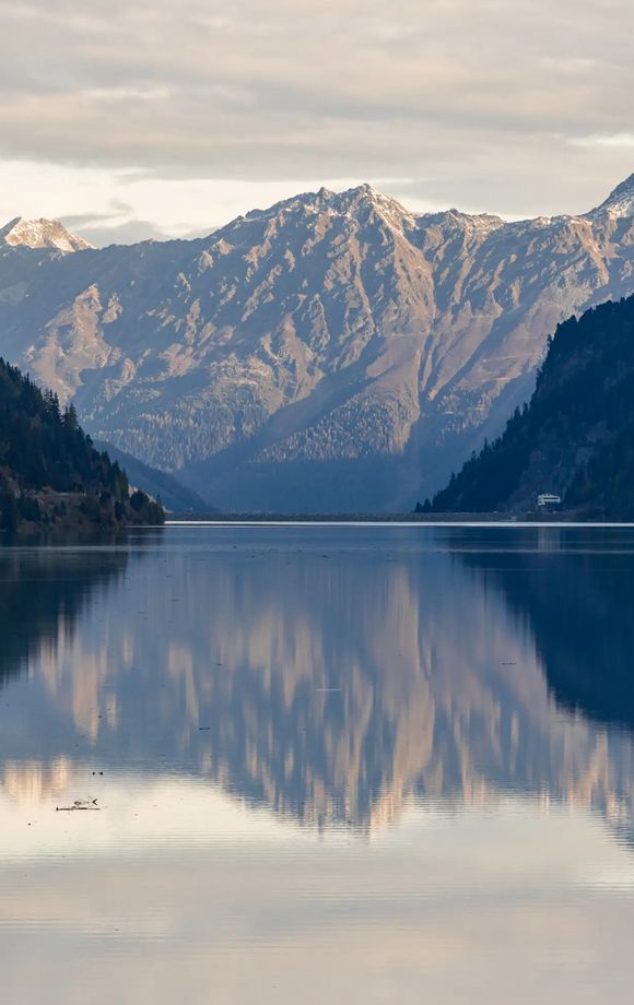 Spiegelglatte Wasseroberfläche des Gepatsch-Stausees im Kaunertal, eingerahmt von bewaldeten Hängen. Die Berggipfel im Hintergrund spiegeln sich stimmungsvoll im Wasser.