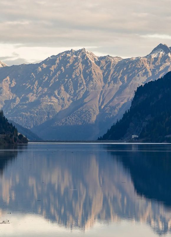 Spiegelglatte Wasseroberfläche des Gepatsch-Stausees im Kaunertal, eingerahmt von bewaldeten Hängen. Die Berggipfel im Hintergrund spiegeln sich stimmungsvoll im Wasser.